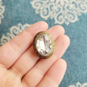 Puede incluir: Una gema de cristal transparente de forma ovalada engastada en un marco de metal color bronce. El marco presenta un diseño de greca alrededor de la gema. La gema está facetada, reflejando la luz. Es probable que sea un botón o un elemento decorativo.