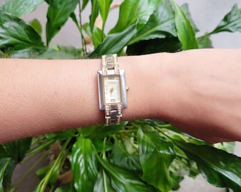 May include: A silver and gold tone watch with a square face and a crystal-encrusted band. The watch face has a white background with black Roman numeral hour markers and black hands.