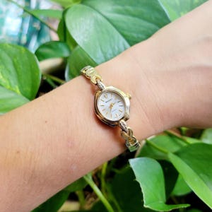 May include: A gold-toned SHARP quartz watch with a white face and gold link bracelet. The watch has a small, oval-shaped face with gold hands and markers. The bracelet is made of interlocking gold links. The watch is worn on a wrist.