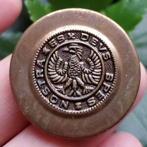 Könnte beinhalten: Ein goldfarbener Metallknopf mit einem erhabenen Design eines Adlers mit ausgebreiteten Flügeln. Der Knopf ist mit den Worten "STRAS DESX DEVS" um den Adler herum beschriftet.