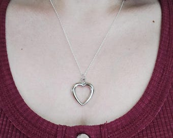 May include: A silver-colored heart pendant necklace. The pendant is a hollow heart shape, suspended from a delicate chain. The necklace is displayed against a maroon ribbed top.