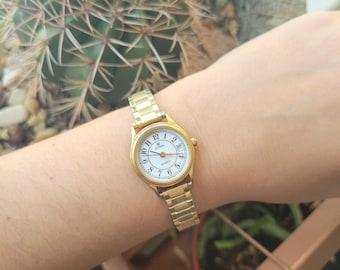 Könnte beinhalten: Eine goldfarbene Armbanduhr mit weißem Zifferblatt und schwarzen römischen Ziffern. Die Uhr hat ein goldfarbenes Metallband.