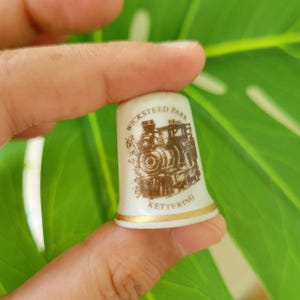 May include: White ceramic thimble with a gold trim. The thimble features a brown illustration of a train with the text "Wicksteed Park Kettering" on it.