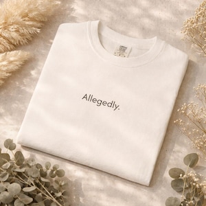 Peut inclure: Un t-shirt blanc à col rond avec le mot "Allegedly." imprimé en noir. Le t-shirt est plié proprement sur une surface de ton neutre, entouré de plantes séchées.