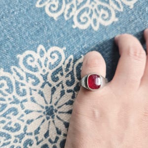 May include: A silver ring with a rectangular, deep red gemstone. The ring is worn on a finger, showcasing the polished stone's vibrant color and the ring's simple, classic design. The ring is set against a blue and white patterned background.