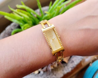 May include: A gold-tone watch with a rectangular face and a crystal-encrusted bezel. The watch has a gold-tone band with a clasp closure.