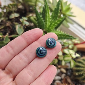 May include: Two round, teal and dark blue earrings with a concentric circle design. The earrings are set against a background of green plants and are held in a person's hand. The earrings are small and appear to be made of a glossy material.