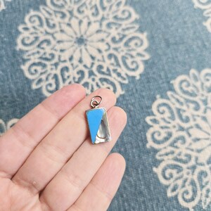 Puede incluir: Un colgante rectangular de plata con un diseño de esmalte azul claro y una incrustación nacarada. El colgante se sostiene en una mano sobre un fondo azul estampado. El colgante tiene un pequeño lazo en la parte superior para una cadena.