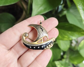 May include: A vintage brooch in the shape of a Viking ship. The brooch features gold-tone metal with black enamel accents and small, clear rhinestones. The sail has a decorative design. A classic accessory.
