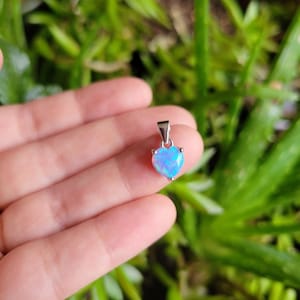 May include: A heart-shaped pendant with a vibrant blue opal stone. The pendant has a silver-colored bail and is set against a blurred green background. The opal has a shimmering, iridescent quality.