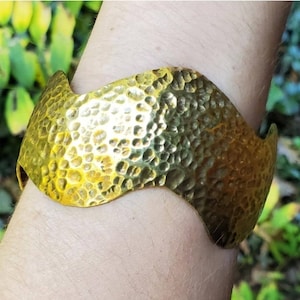 Puede incluir: Un brazalete de metal martillado de color dorado con un diseño ondulado. El brazalete se lleva en la muñeca.