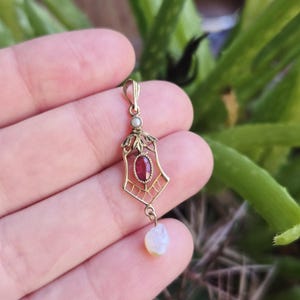 Puede incluir: Un colgante dorado con una piedra preciosa roja y una pequeña perla. El colgante tiene un diseño ornamentado, de inspiración Art Déco, con un patrón geométrico y detalles de hojas. Una perla en forma de lágrima cuelga de la parte inferior.