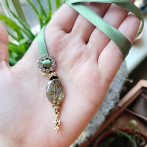 May include: A delicate green satin ribbon necklace with a gold-toned pendant featuring a clear oval resin charm with dried botanicals and a small rhinestone embellishment.