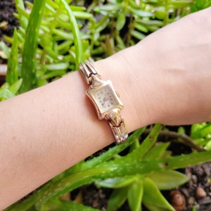 Könnte beinhalten: Eine goldfarbene Armbanduhr mit quadratischem Zifferblatt und einem zarten Gliederarmband. Das Zifferblatt der Uhr hat schwarze römische Ziffern für die Stunden und einen kleinen Sekundenzeiger.