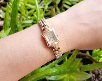 May include: A gold-toned wristwatch with a square face and a delicate, linked band. The watch face has black Roman numeral hour markers and a small second hand.