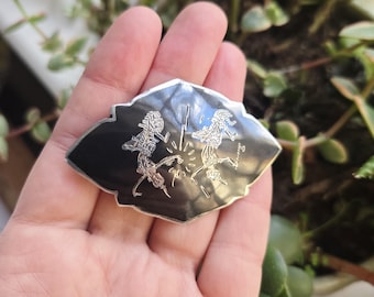 May include: A silver and black brooch with a unique design. The brooch features two stylized figures with a central burst of light. The figures are in a dancing pose. The brooch is held in a hand.