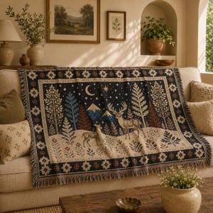 Könnte beinhalten: Eine gewebte Wohndecke mit dunkelblauem Hintergrund und Waldmotiv. Das Design zeigt einen großen Hirsch, ein kleineres Reh, schneebedeckte Berge, Tannenbäume, eine Mondsichel und Sterne. Der Rand ist mit einem floralen Muster verziert.