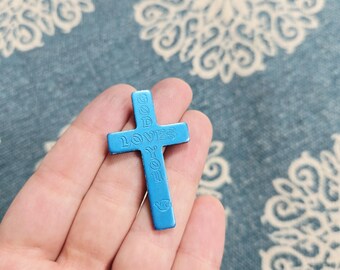 May include: A light blue cross with the words "GOD LOVES YOU" embossed on it. The cross is held in a hand against a patterned blue background. The cross is a religious item.
