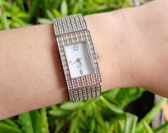 May include: A silver watch with a square face and a crystal-encrusted band. The watch face has a white background with black Roman numeral hour markers and black hands. The watch face also has the word "QUARTZ" printed on it.