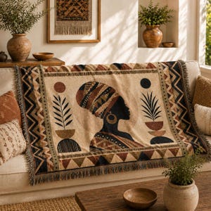 May include: A woven throw blanket featuring a profile illustration of a person wearing a patterned headwrap. The design includes geometric borders, stylized plant motifs in pots, and circular shapes in shades of brown, black, and cream.