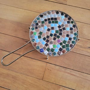 May include: A round, mosaic-tiled trivet with a metal handle. The trivet is made of small, colorful tiles in shades of blue, green, brown, and white. The trivet is on a wooden surface.