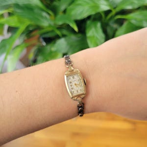 May include: A gold-toned wristwatch with a white face and black Roman numeral hour markers. The watch has a rectangular face and a metal band.