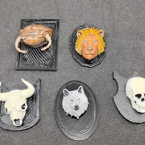 May include: A selection of miniature animal head wall mounts, including a crab, lion, bull, wolf, and skull. Each is mounted on a black plaque. The lion has a yellow mane, and the wolf is grey. Measurements are in centimetres.