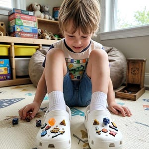 Könnte beinhalten: Ein Paar weiße Gummischuhe, verziert mit verschiedenen kleinen, bunten Fantasy-Anhängern wie Miniaturschwertern, Schatztruhen und mehrseitigen Würfeln. Die Schuhe werden mit weißen Socken getragen und haben Belüftungslöcher auf der Oberseite.
