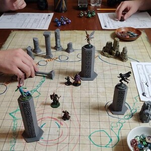 May include: A tabletop role-playing game setup on a grid map featuring gray 3D-printed vertical height trackers with numerical markings, various painted miniature figures, stone-like terrain pieces, and a bowl filled with colorful glass beads.