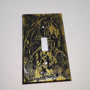May include: Black and gold light switch cover featuring a detailed dragon design. The dragon is outlined in gold against a textured black background. The switch itself is white.