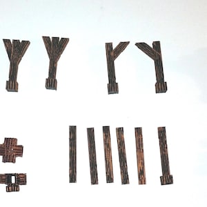 May include: A collection of small, dark brown wooden pieces, likely for a model or craft project. The pieces include cross-shaped components, vertical supports, and angled beams. The wood has a visible grain, suggesting a natural finish. The arrangement suggests a construction theme.