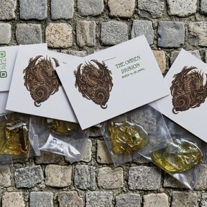 Könnte beinhalten: Mehrere kleine, transparente Plastiktüten mit bernsteinfarbenen, unregelmäßig geformten Objekten werden gezeigt. Jede Tüte ist mit einer weißen Karte mit einer braunen Drachenillustration und dem Text "THE OMEN DRAGON" versehen. Ein QR-Code ist ebenfalls sichtbar.