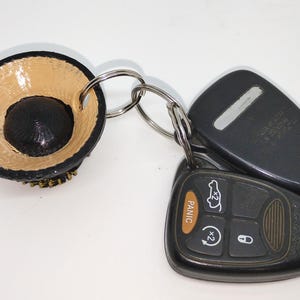 Puede incluir: Un llavero hecho a mano con un pequeño dije redondo en forma de cuenco negro y beige y un anillo de metal, sujeto a un juego de llaves de coche. Las llaves son negras con detalles plateados y un botón de pánico.