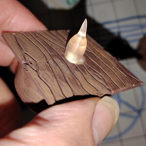 May include: A miniature brown wooden structure with a clear, pointed object protruding from a hole. The wood has a textured, grooved surface. The object appears to be lit from within, creating a soft glow.