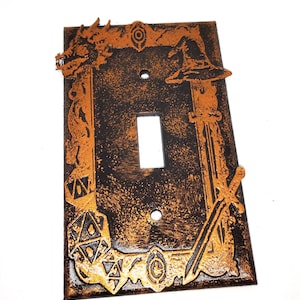 May include: A decorative light switch cover with a fantasy theme. The cover is black with bronze-colored accents. It features a dragon, wizard hat, sword, and dice. The switch opening is white.