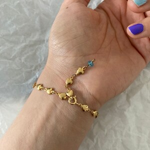 Può includere: Un braccialetto a catena dorata con un design a forma di cuore e una piccola perla blu. Il braccialetto è al polso.