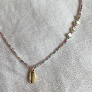 Könnte beinhalten: Eine zarte Halskette aus bunten Perlen und einem Muschelanhänger. Die Halskette hat auf einer Seite eine Reihe kleiner weißer Muschelperlen.