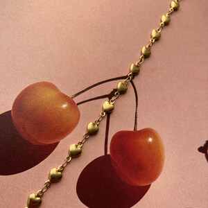 May include: A gold chain bracelet with a series of small heart charms. The bracelet is lying on a pink surface with two red cherries.
