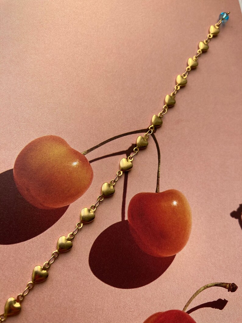 May include: A gold chain bracelet with a series of small heart charms. The bracelet is lying on a pink surface with two red cherries.