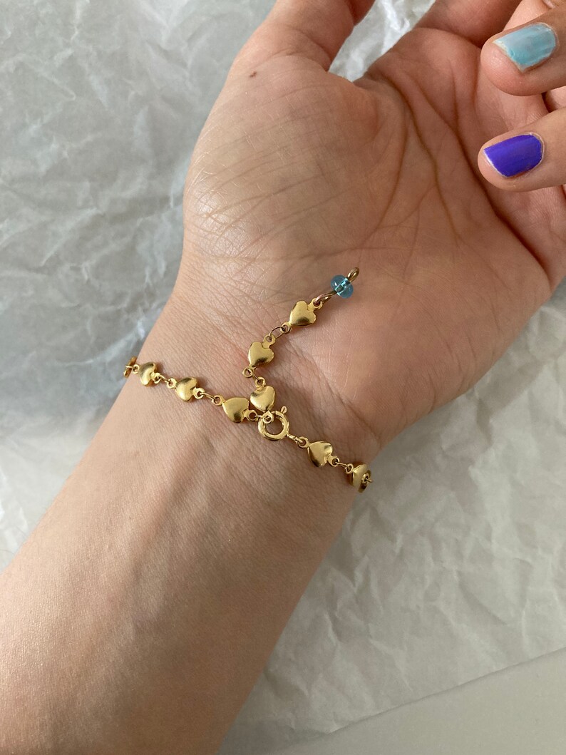 May include: A gold chain bracelet with a heart-shaped design and a small blue bead. The bracelet is on a person's wrist.