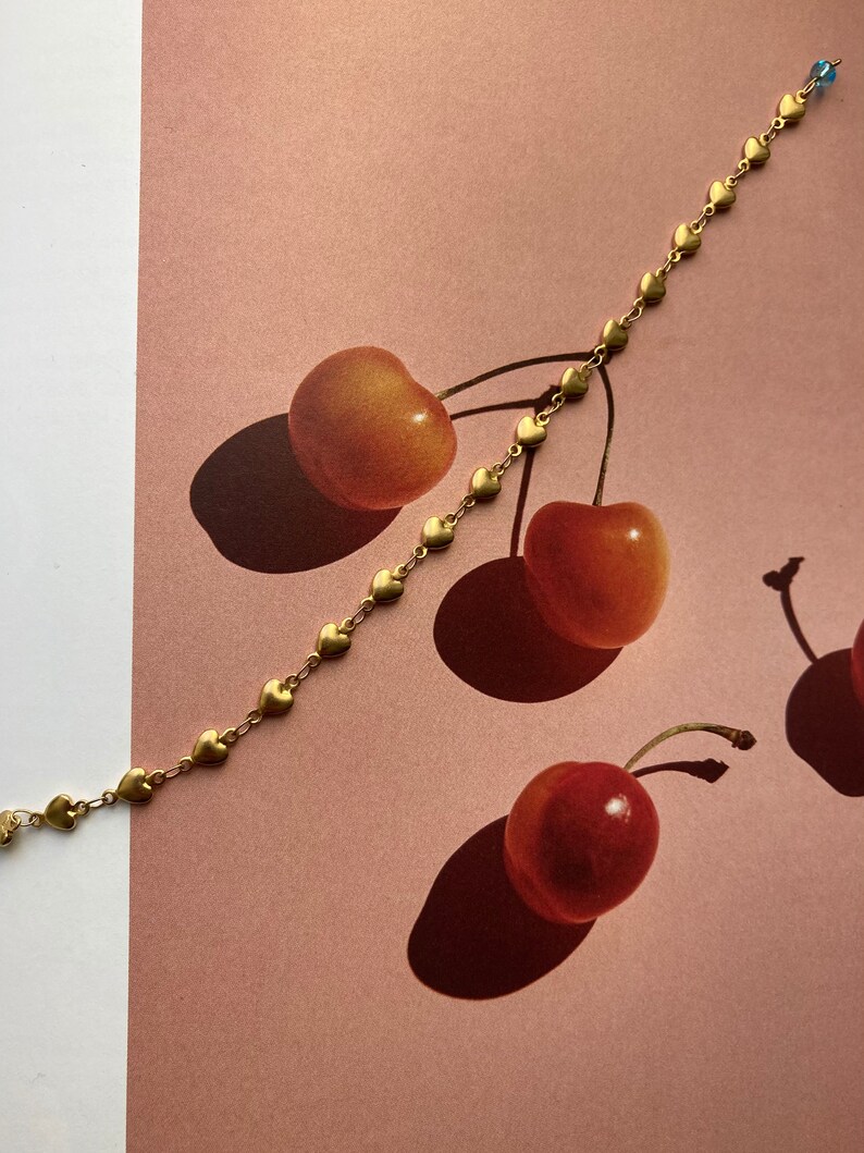 May include: A gold chain necklace with small heart-shaped links. The necklace is laying on a pink surface with three red cherries.