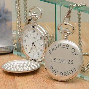 May include: Silver pocket watch with a white face and black numerals. The watch is open, revealing the time. The back of the watch reads "FATHER OF THE BRIDE 18.04.26". The watch is on a silver chain.