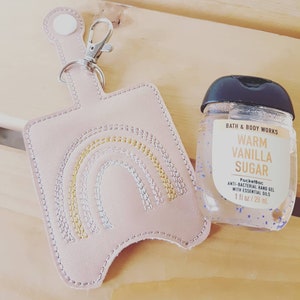 Könnte beinhalten: Ein rosa Leder-Schlüsselanhänger mit einem Regenbogen-Design und eine kleine Flasche Bath & Body Works Warm Vanilla Sugar PocketBac Antibakterielles Handgel mit ätherischen Ölen. Die Flasche ist 1 fl oz / 28 ml.