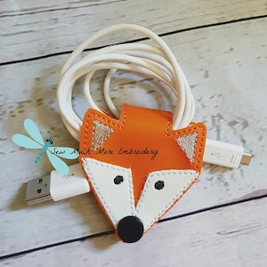 May include: Orange fox-shaped cable organizer with white and brown embroidery details. The organizer is holding a white charging cable.
