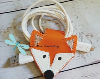 May include: Orange fox-shaped cable organizer with white and brown embroidery details. The organizer is holding a white charging cable.