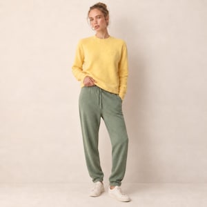 Op de afbeelding: Een okergele trui met lange mouwen en ronde hals gecombineerd met een saliegroene joggingbroek met een elastische tailleband met trekkoord en boorden bij de enkels, gedragen met witte sneakers. De outfit bestaat uit een zachte gebreide set.