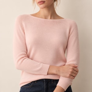May include: A light pink long-sleeve sweater featuring a wide boat neckline and ribbed cuffs. The knit garment has a smooth texture and raglan sleeves, providing a clean and simple look that pairs well with dark blue denim jeans.