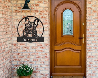 May include: Black metal wall art featuring a French Bulldog within a circular frame. The dog's name, "WINSTON," is displayed on a rectangular plaque below. The artwork is mounted on a brick wall near a wooden door.