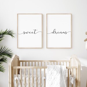 Puede incluir: Dos impresiones enmarcadas con las palabras "sweet dreams" en una fuente cursiva. Las impresiones están colgadas en una pared blanca sobre una cuna de madera.