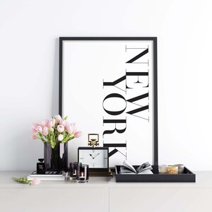 K&ouml;nnte beinhalten: Ein schwarzer gerahmter Druck mit dem Wort "NEW YORK" vertikal in Wei&szlig; geschrieben. Der Druck befindet sich auf einem wei&szlig;en Regal mit einem schwarzen Tablett, einer goldenen Uhr, einer Vase mit rosa Tulpen und zwei Kerzenhaltern.